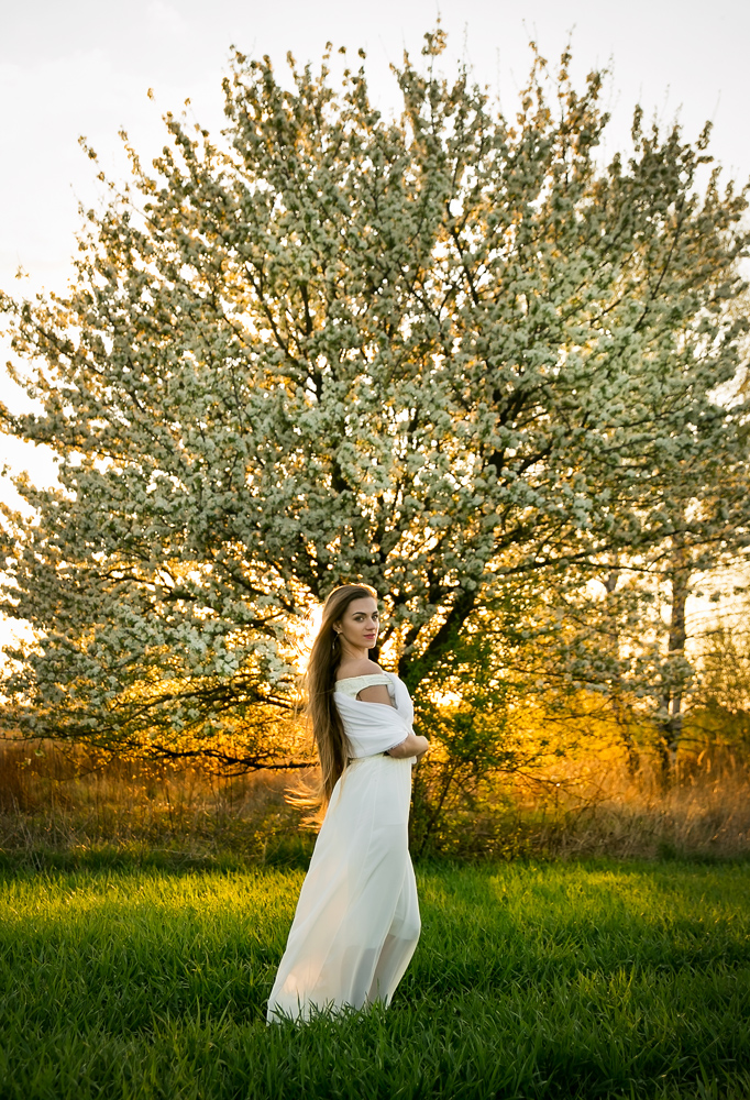 Piękna kobieta w białej sukience na łące podczas kobiecej sesji w plenerze o zachodzie słońca.